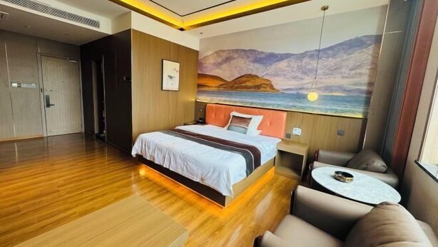 A modern hotel room with a wooden floor, a bed with a white and brown bedspread, a small round table, and a painting of a sunset over mountains.