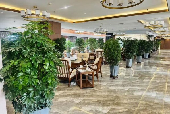 Un amplio vestíbulo de hotel con piso de mármol, una zona de comedor con sillas y mesas de madera, y varias plantas en macetas.
