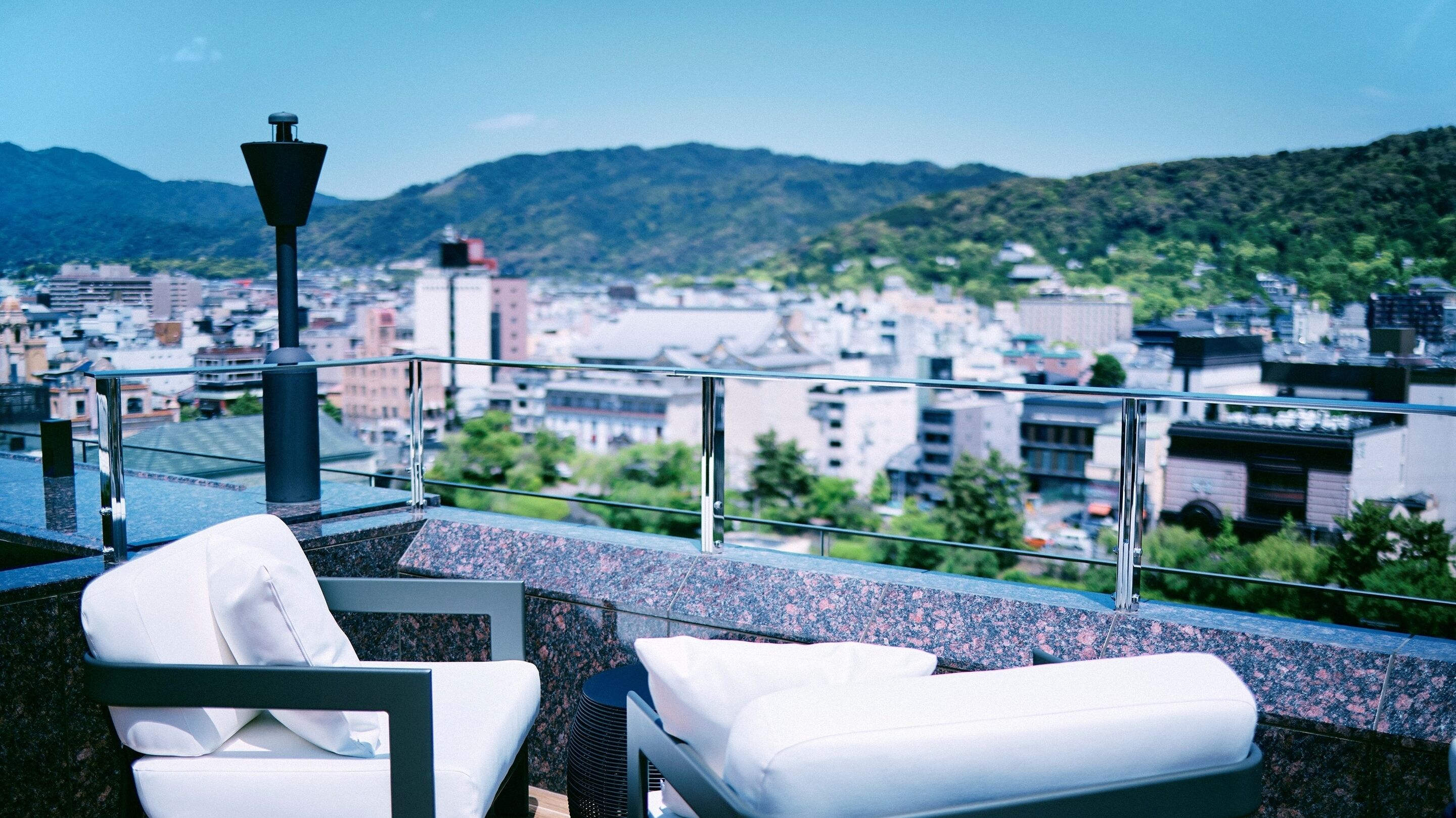 A rooftop terrace with two white lounge chairs, a metal railing, and a view of a cityscape with buildings and mountains in the background.
