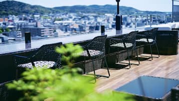 A rooftop terrace with wicker chairs and a table, overlooking a cityscape with buildings and hills.