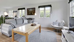 Un salon spacieux avec une table basse en bois, un canapé gris et un fauteuil blanc. Un tableau orne le mur, une fenêtre offre une vue sur la verdure, et un téléviseur est posé sur un meuble blanc.