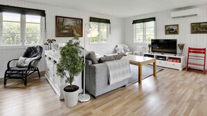 Un salon chaleureux avec un canapé, une table à manger, un téléviseur et un fauteuil rouge. Il y a des fenêtres avec des rideaux et une plante en pot.