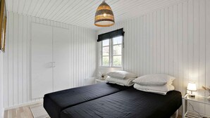 Une chambre avec un grand lit, une fenêtre ornée d’une lambrequin noire, un luminaire suspendu en osier et des murs recouverts de panneaux blancs.