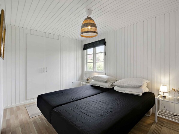 Une chambre avec un grand lit, une fenĂȘtre ornĂ©e dâune lambrequin noire, un luminaire suspendu en osier et des murs recouverts de panneaux blancs.