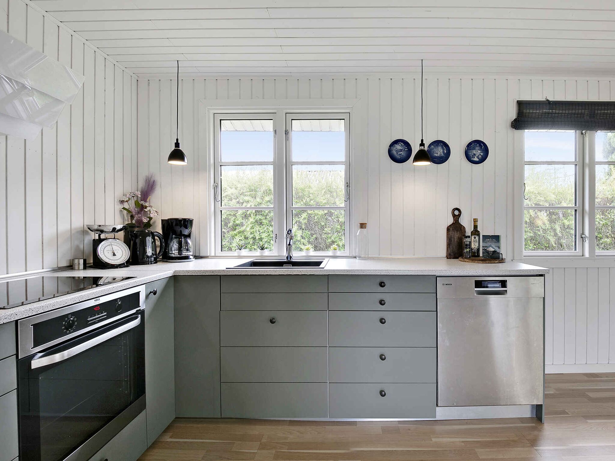 A modern kitchen with a large island, built-in oven, and a dishwasher. There are two windows providing natural light and a view of greenery outside.