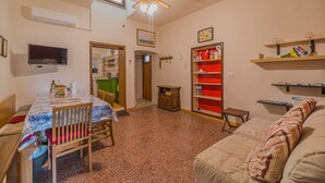 A cozy living room with a sofa, a dining table with chairs, a TV mounted on the wall, a ceiling fan, and a kitchen area visible through an open door. - BILOCALE NEL CENTRO DI PORTOFERRAIO (Portoferraio)