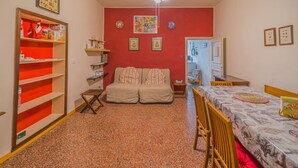A compact room with a red wall, a ceiling fan, a sofa, a dining table, and a small kitchen area. - BILOCALE NEL CENTRO DI PORTOFERRAIO (Portoferraio)