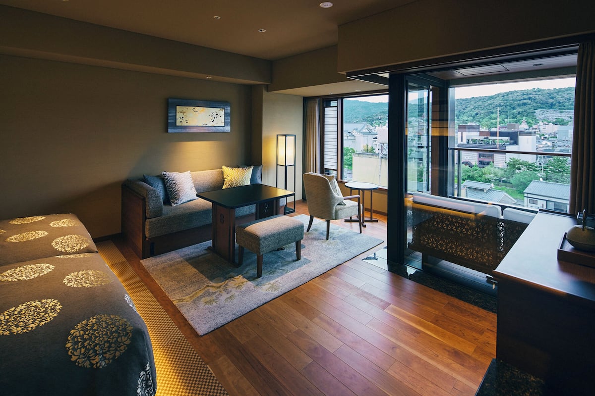 Une chambre d’hôtel moderne équipée d’un canapé, d’une petite table, d’une chaise et d’un balcon offrant une vue sur la ville et les collines.