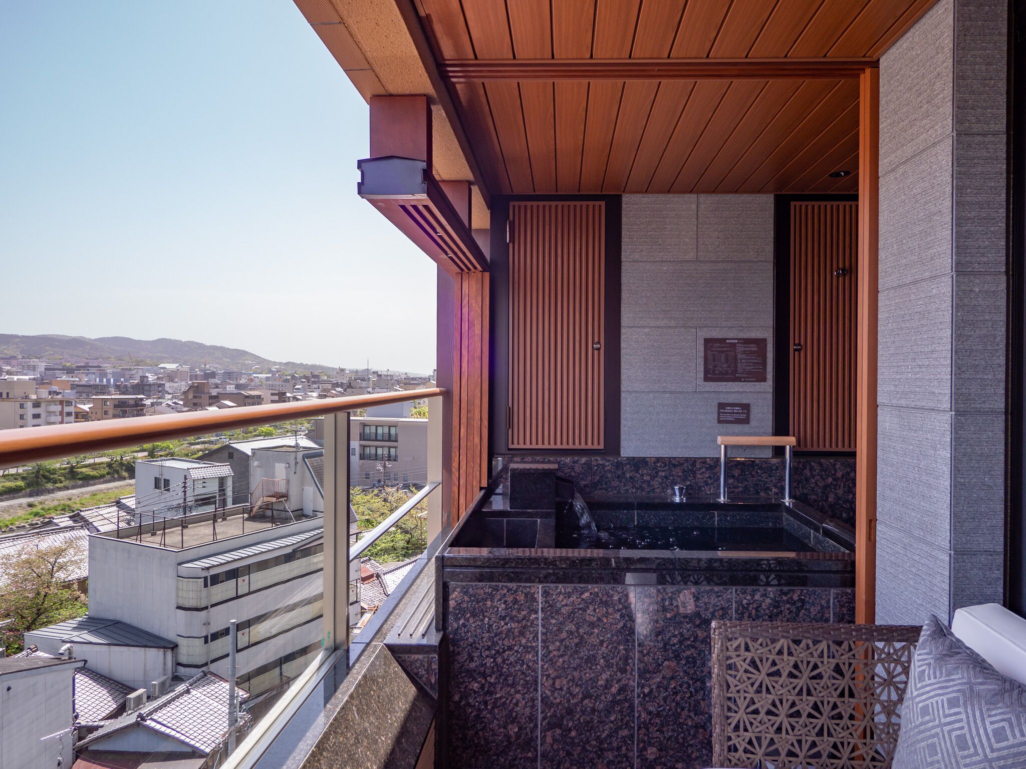 A modern balcony with a view of a cityscape, a built-in grill, and a seating area with a patterned cushion.