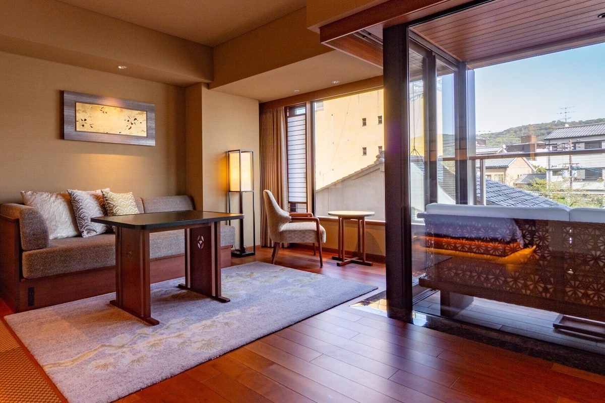 Une chambre d’hôtel moderne équipée d’un canapé, d’une petite table, d’une chaise, d’un lampadaire et d’une grande fenêtre donnant sur la ville.