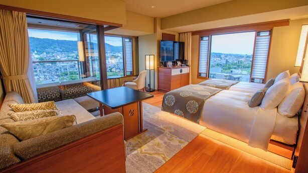 A hotel room with a sofa, a bed, a desk, and a TV. The room has large windows with a view of a city and mountains. - Soraniwa Terrace Kyoto Bettei (Kyoto)