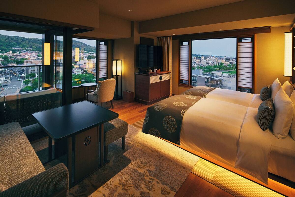 Une chambre d’hôtel avec un grand lit, un bureau, une chaise et une télévision. La chambre dispose de grandes fenêtres donnant sur la ville.