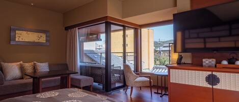 A hotel room with a sofa, a small table, a TV, and a balcony with a view of the city.