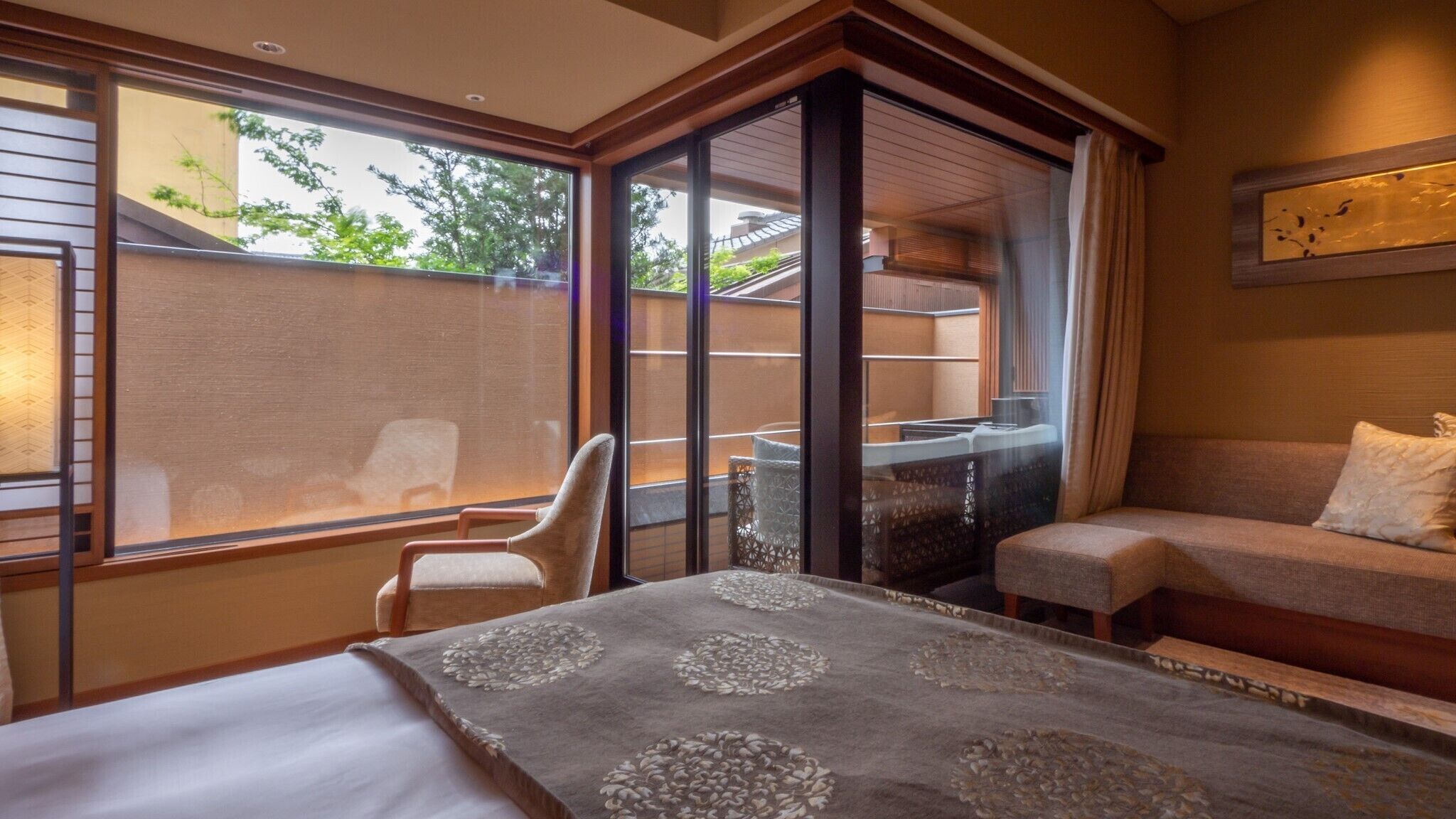 A hotel room with a bed, a seating area, and a balcony with a view of greenery.
