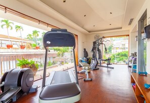 Un gimnasio con caminadora, elíptica y máquinas de pesas. Tiene ventanas amplias con vistas a palmeras y un edificio.