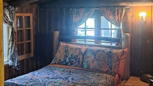 A rustic wooden cabin bedroom with a bed, a wooden headboard, a window with curtains, a wall-mounted lamp, and a fish decoration on the wall.