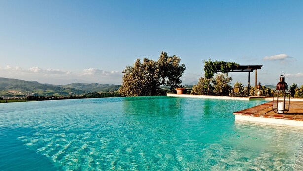 A clear blue infinity pool with a wooden deck, surrounded by lush greenery and a pergola. - SOTTO COLLE - COTTAGE (Montelupo Fiorentino)