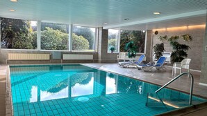 Une piscine intérieure à l’eau d’un bleu cristallin, entourée de chaises longues et dotée de grandes fenêtres offrant une vue sur la verdure.