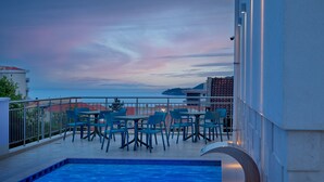A rooftop pool area with blue chairs and tables, a pool, and a view of the sea and buildings. - Larimar (Becici)