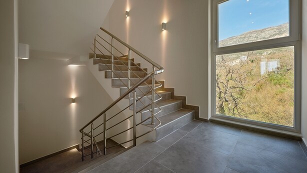A modern staircase with metal railings and built-in lighting, leading to a large window with a view of a hilly landscape. - Larimar (Becici)