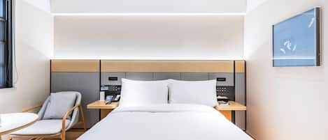 A modern hotel room with a large bed, a bedside table with a phone and a digital device, a chair, and a small round table.
