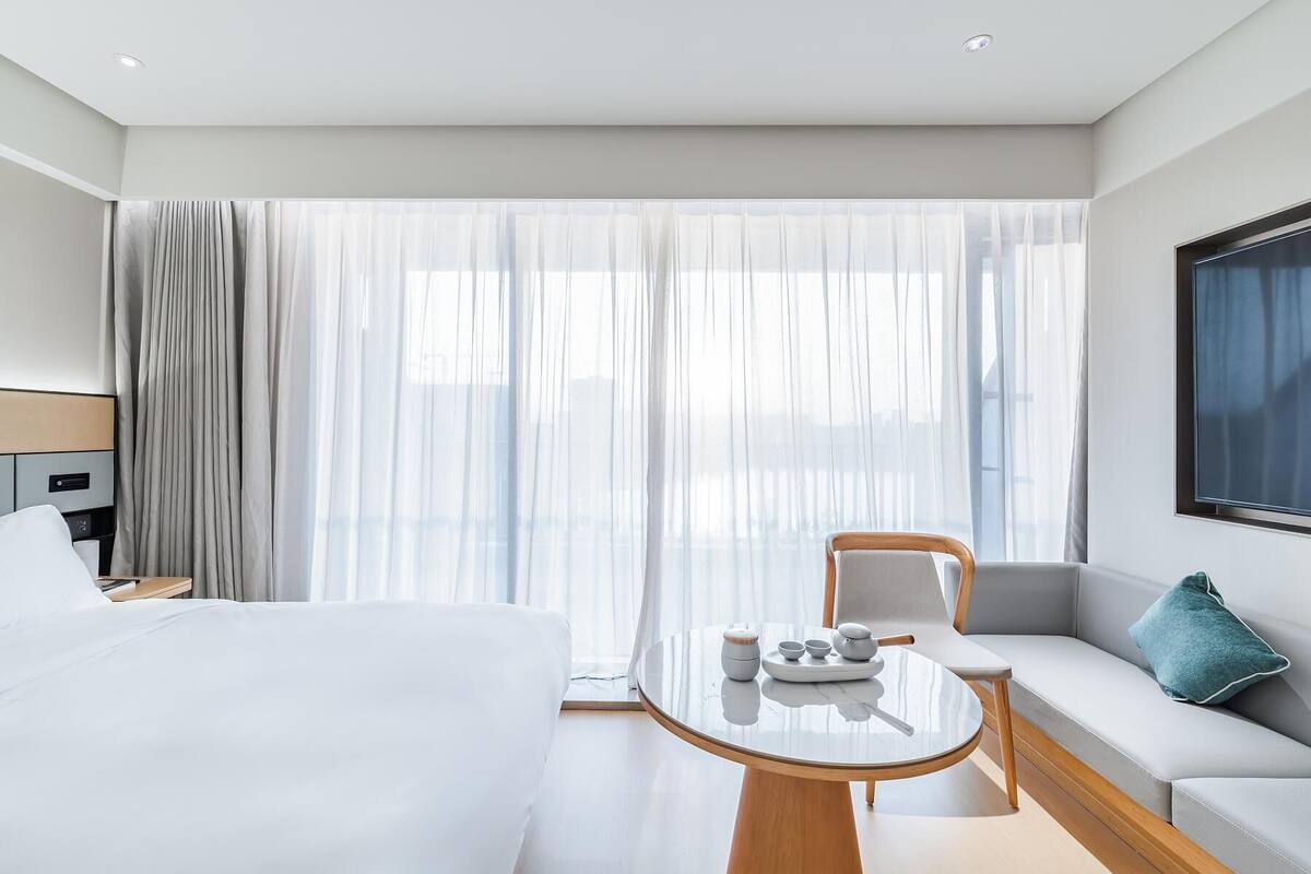Una habitación de hotel con cama, sofá, mesa con bandeja de té y ventana con cortinas translúcidas.
