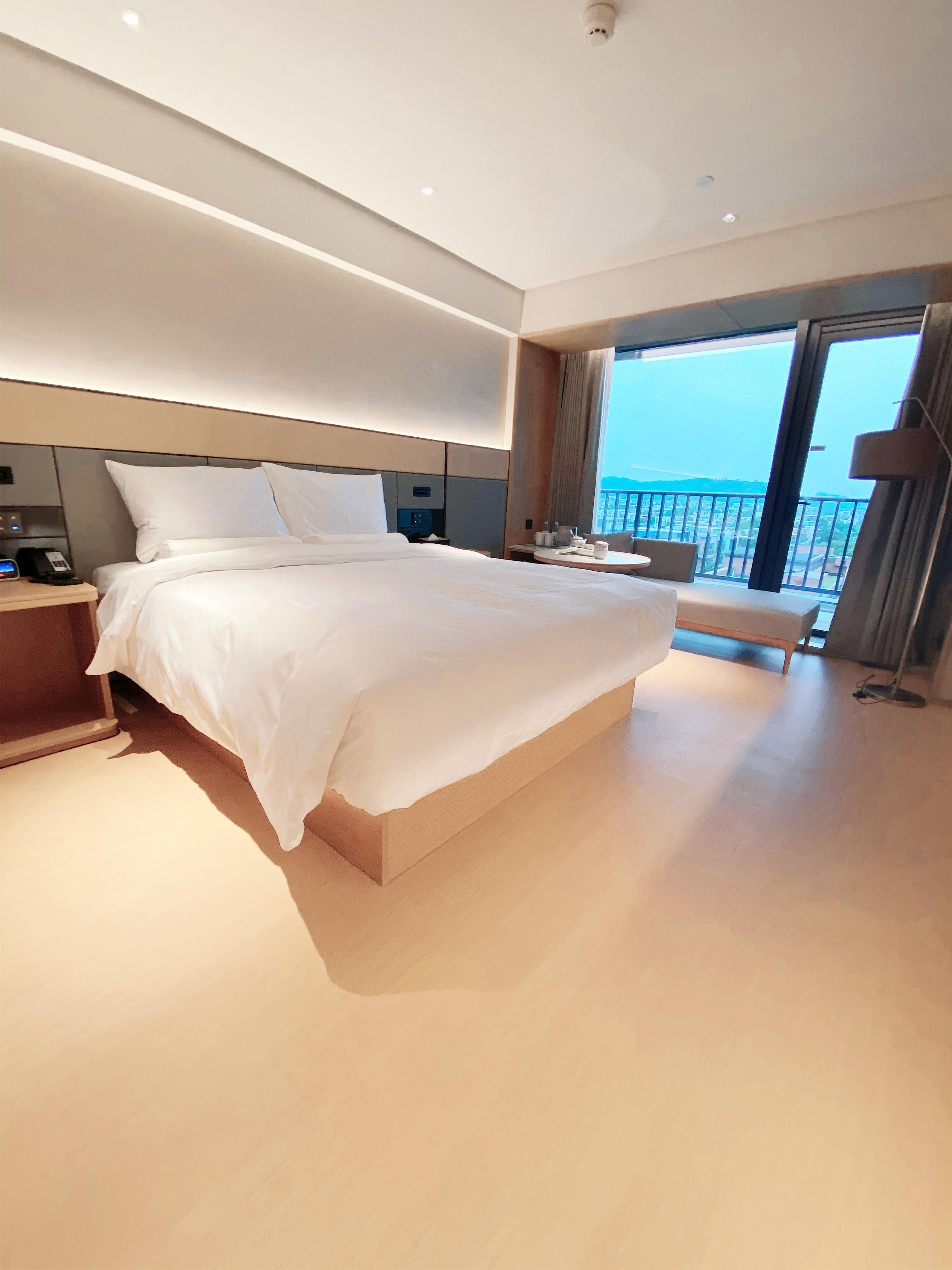 A modern hotel room with a large bed, bedside table, and a view of the city through the balcony.