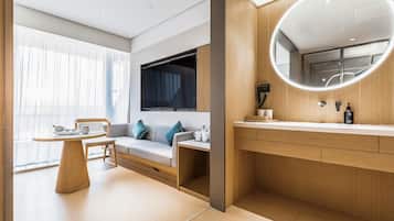Una habitación de hotel moderna con un sofá, una pequeña mesa, un televisor, un espejo redondo y un tocador de madera.