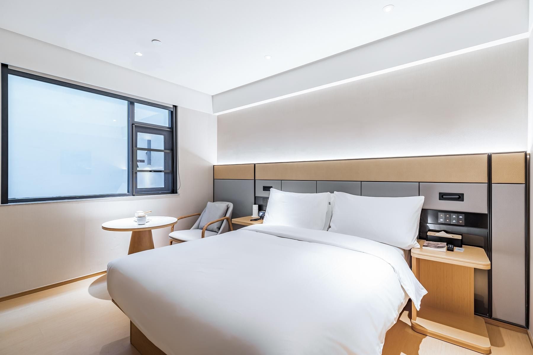 A modern hotel room with a large bed, a bedside table, a chair, and a window.