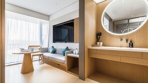A modern hotel room with a sofa, a small table, a TV, a round mirror, and a wooden vanity. - JI Hotel (Nanjing)