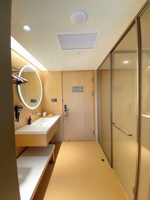 A modern bathroom with a wooden vanity, a mirror with a light, a bench, a shower with a glass door, and a ceiling vent. - JI Hotel (Nanjing)