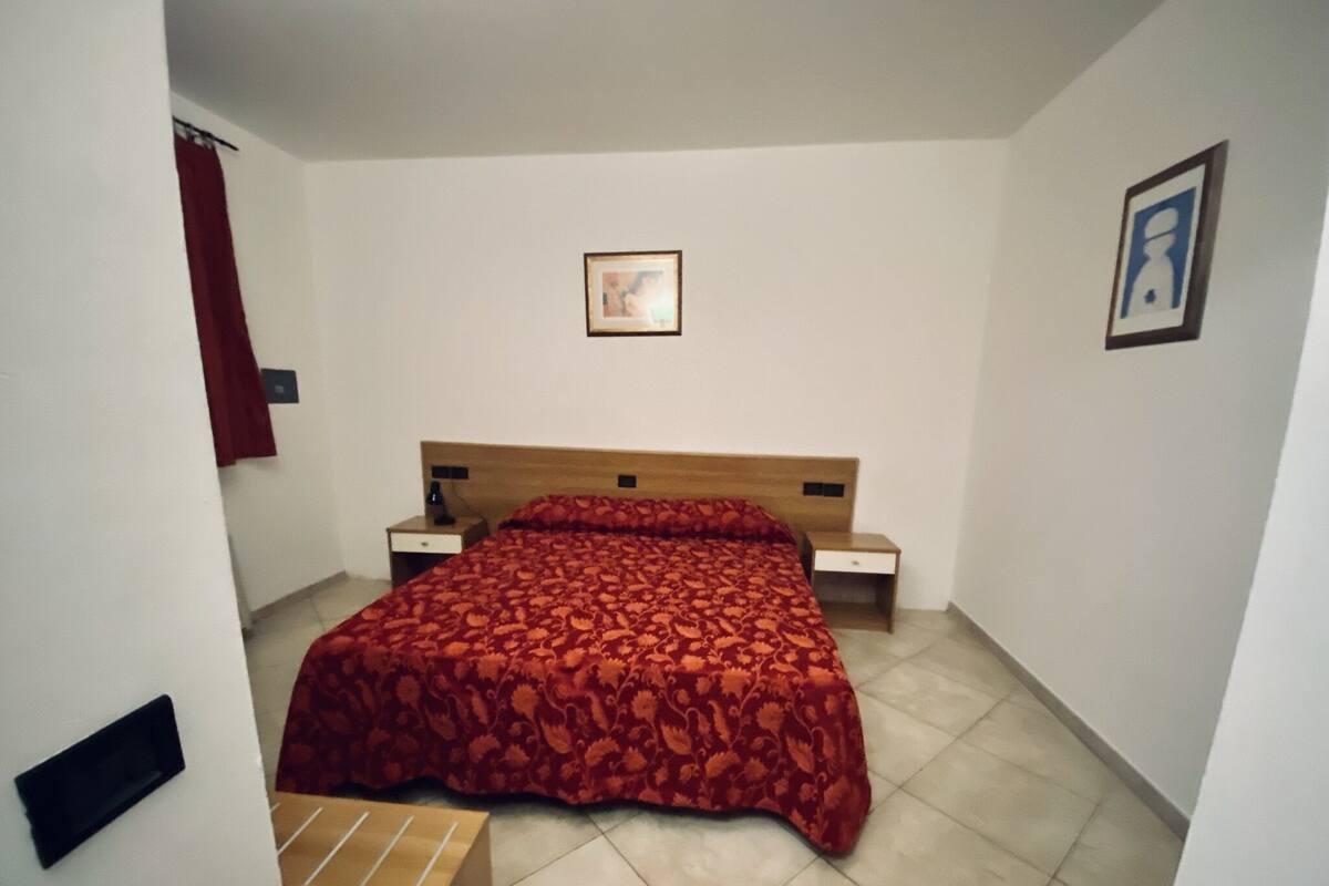 Une chambre à coucher avec un lit, deux tables de chevet, une couvre-lit rouge à motifs, un tableau encadré au mur et un rideau rouge.