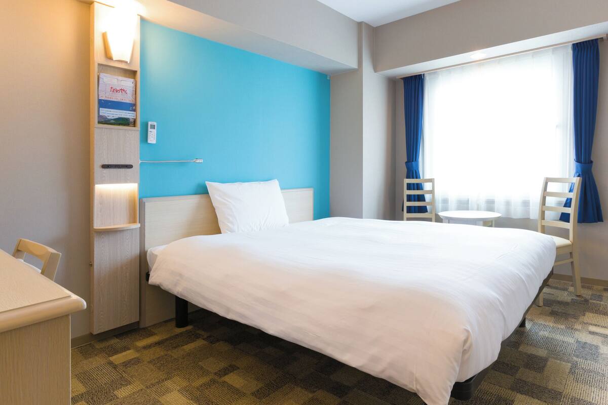 Habitación de hotel con una cama grande, un escritorio, una silla y una mesita pequeña. Cuenta con una ventana con cortinas y un panel de control en la pared.