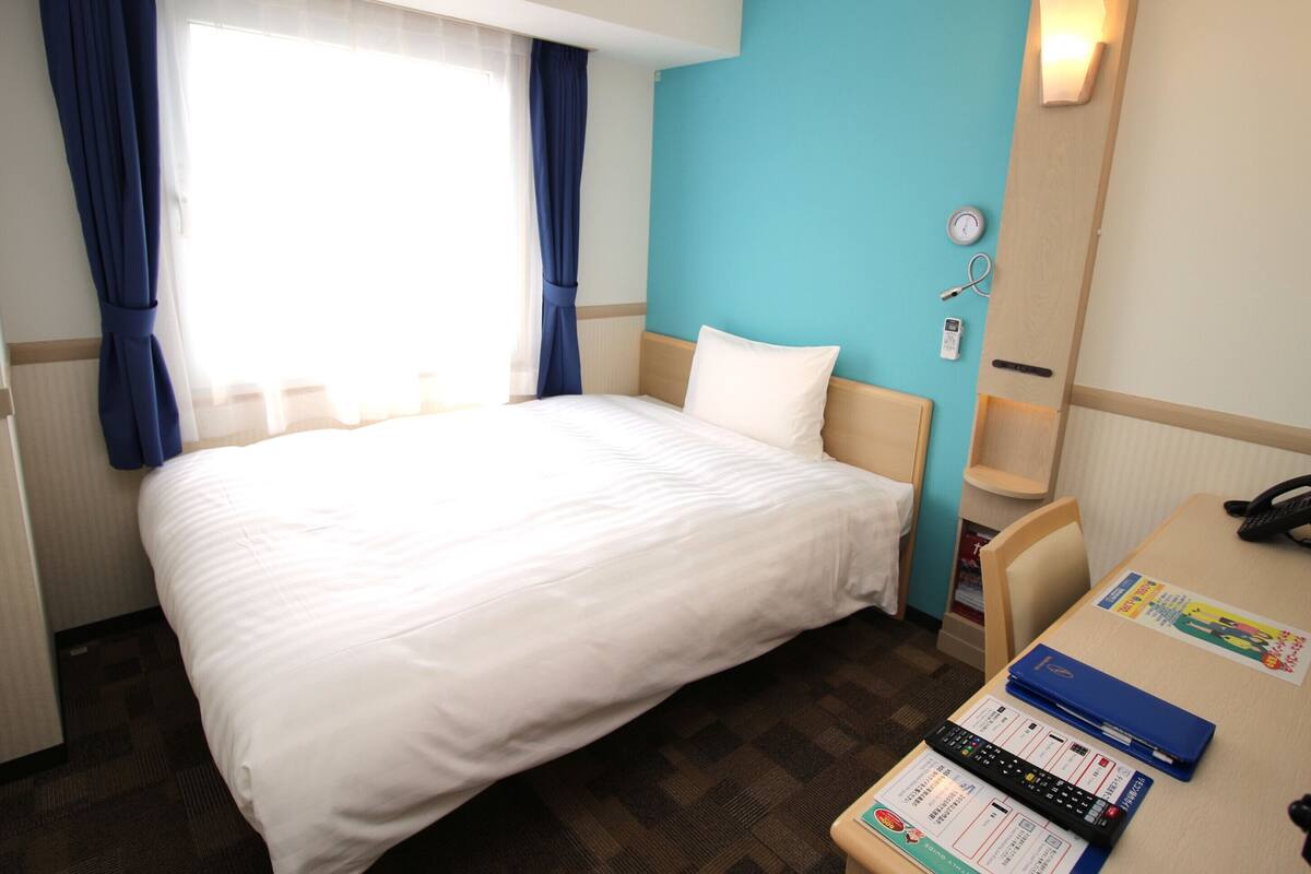 Habitación de hotel con una cama grande, un escritorio con computadora y papeles, un reloj y un teléfono en la pared.