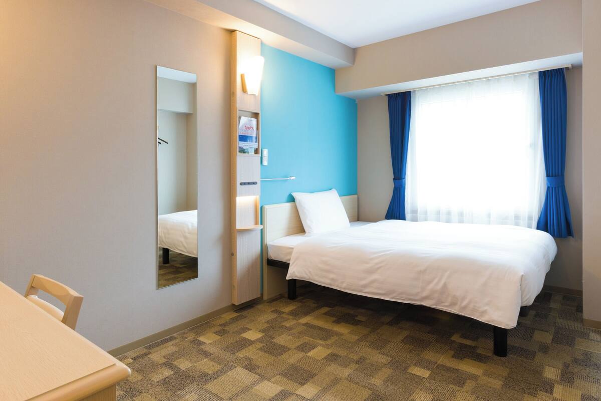 Habitación de hotel con cama, escritorio, silla, espejo, armario y ventana con cortinas.
