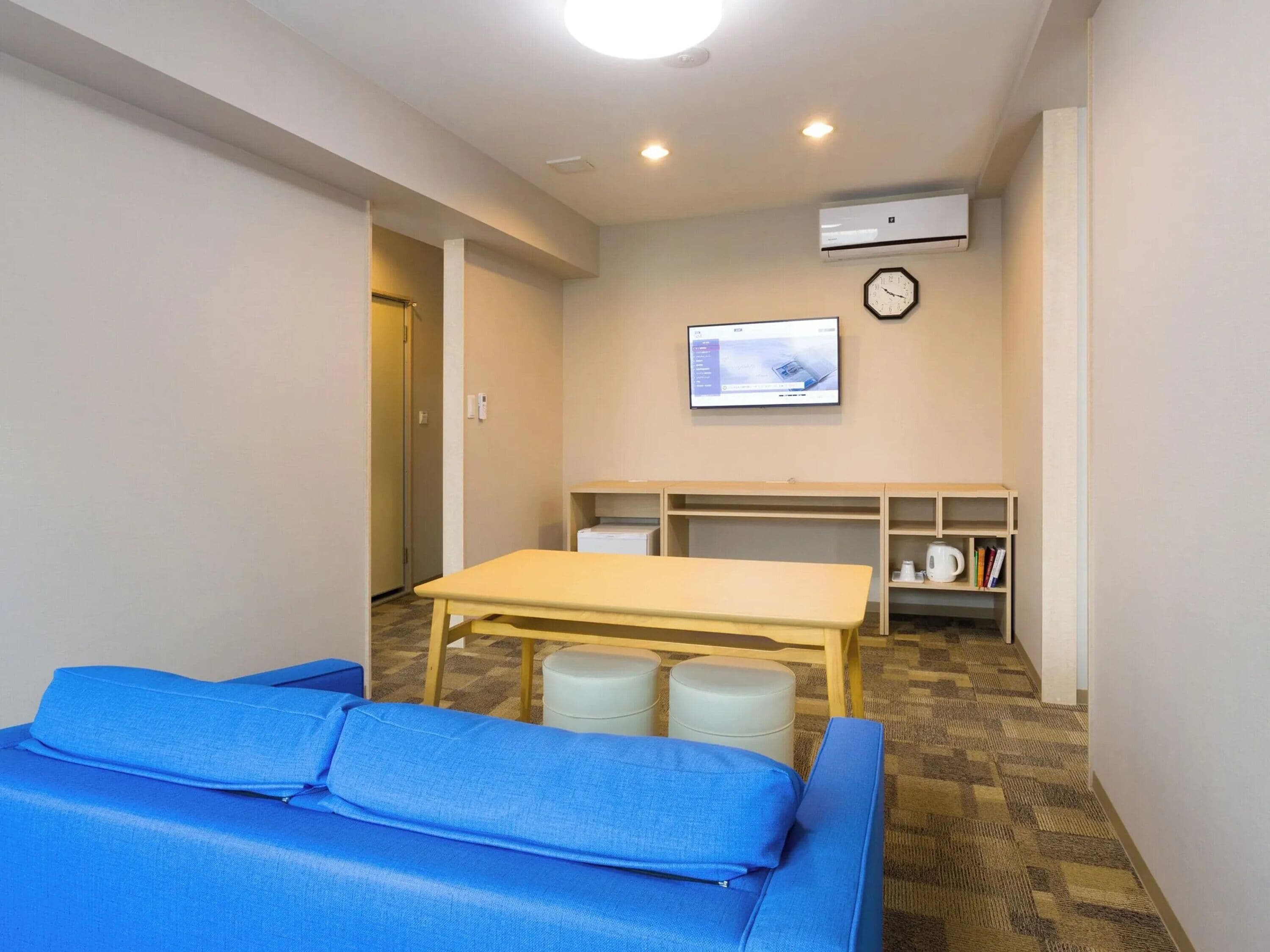 A room with a blue sofa, a wooden table, a television mounted on the wall, and a clock.