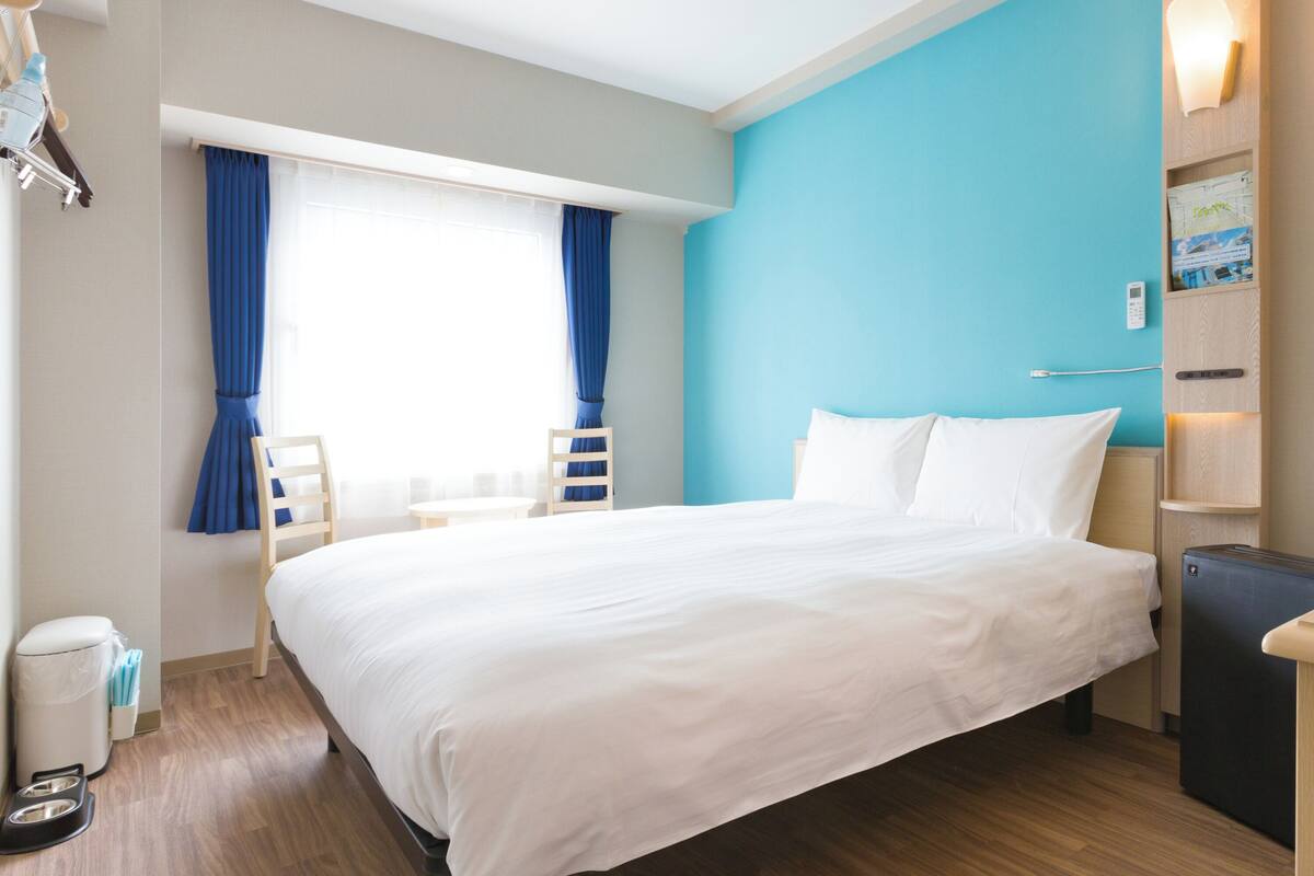 Habitación de hotel con una cama grande, un escritorio, una silla, una mesita, un cubo de basura y una ventana con cortinas.
