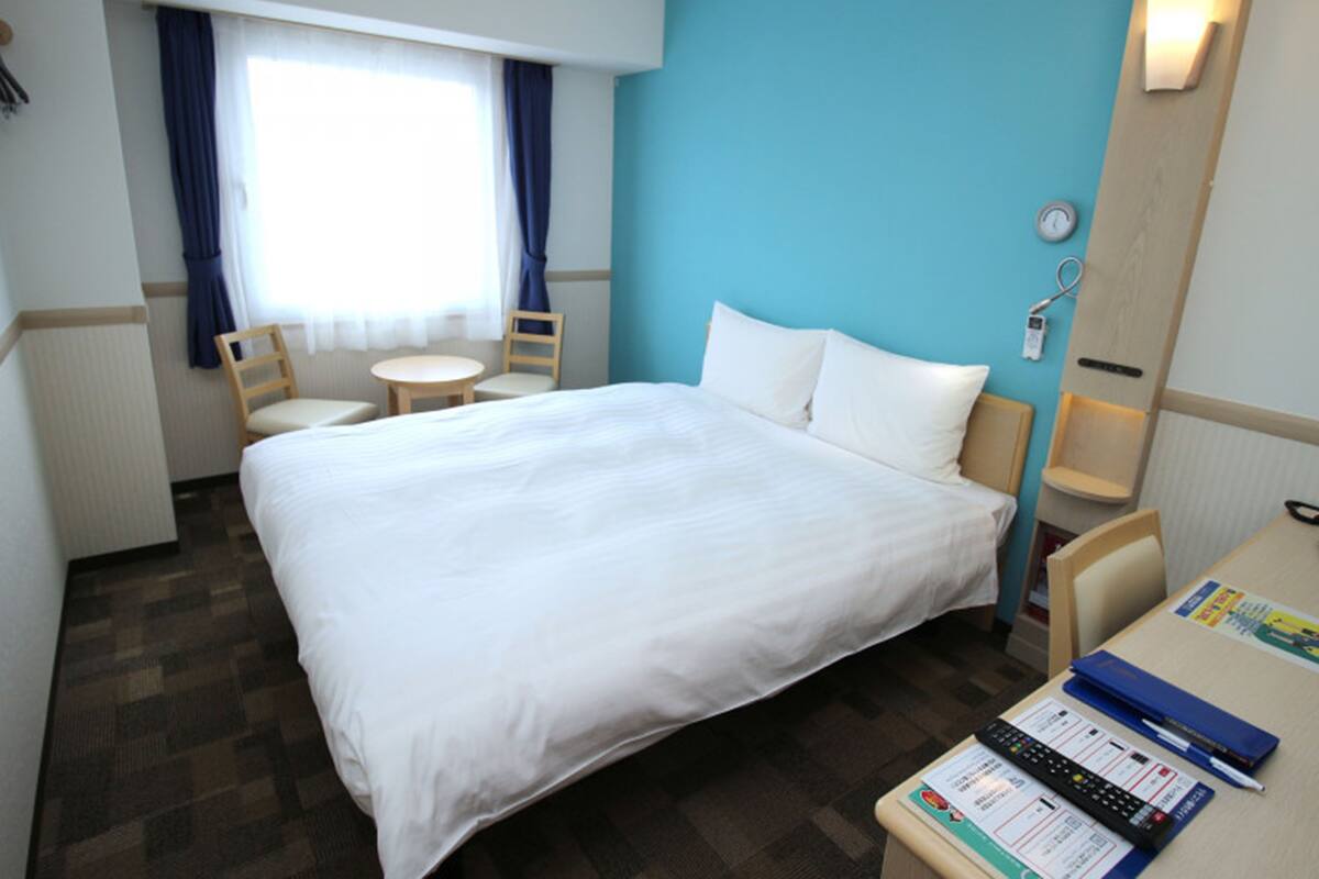 Habitación de hotel con una cama grande, un escritorio con silla, una mesita y un ventanal con cortinas.
