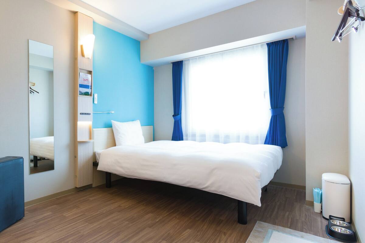 Una habitación de hotel moderna con una cama grande, una mesita de noche, un espejo y una ventana con cortinas azules.