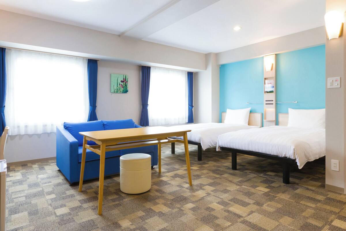 Una habitación de hotel con alfombra a cuadros, un sofá azul, una mesa de madera, dos camas individuales y un otomano pequeño y redondo.