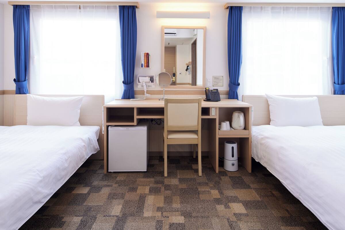 Habitación de hotel con dos camas individuales, un escritorio con silla, un refrigerador pequeño, una cafetera y un espejo.