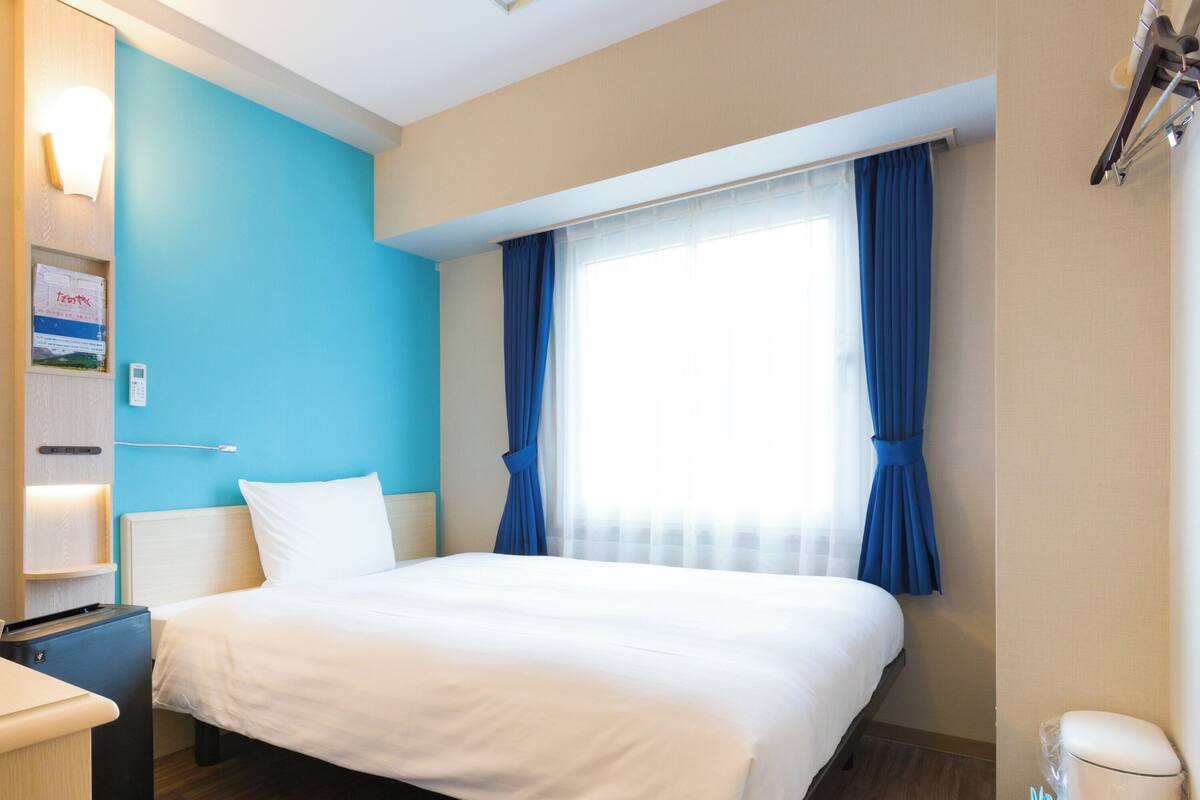 Habitación de hotel con cama, escritorio, mesita de noche, lámpara de pared, ventana con cortinas y teléfono en la pared.