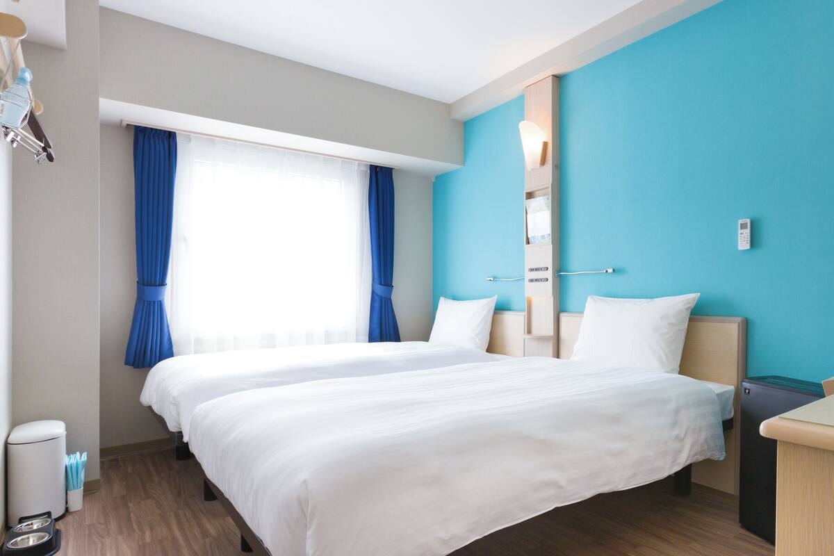 Una habitación de hotel con dos camas, una ventana con cortinas azules, una mesita de noche y un teléfono en la pared.