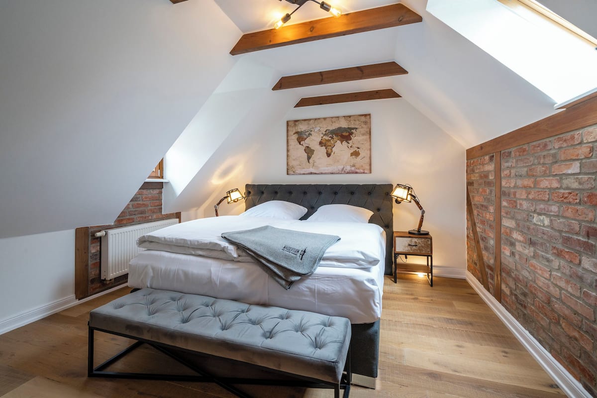 A loft bedroom with a sloped ceiling, exposed wooden beams, a world map on the wall, a bed with white linens, two bedside tables with lamps, and a bench.
