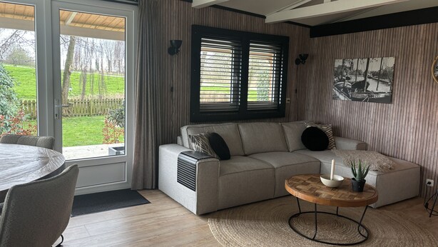 Una sala de estar moderna con un sofá gris, una mesa de centro redonda de madera y vistas a un jardín a través de una puerta corrediza de vidrio.