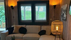 Una sala de estar moderna con un sofá seccional, una mesa de centro redonda y una lámpara de pie. Hay dos ventanas con persianas, una planta en maceta y una alfombra en el suelo.