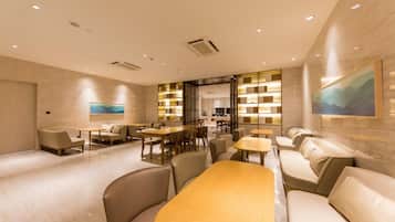 A modern hotel lobby with a wooden reception desk, seating areas, and a view into the dining area.