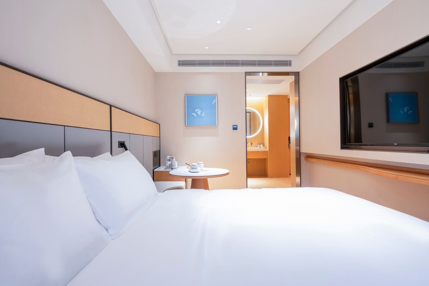 Une chambre d’hôtel moderne avec un grand lit, une table ronde avec une machine à café, et une vue sur la salle de bain grâce à une porte ouverte.