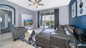 Ein modernes Wohnzimmer mit grauem Sofa, einem Glastisch, einem Deckenventilator und einem Fernseher auf einem weißen Lowboard.