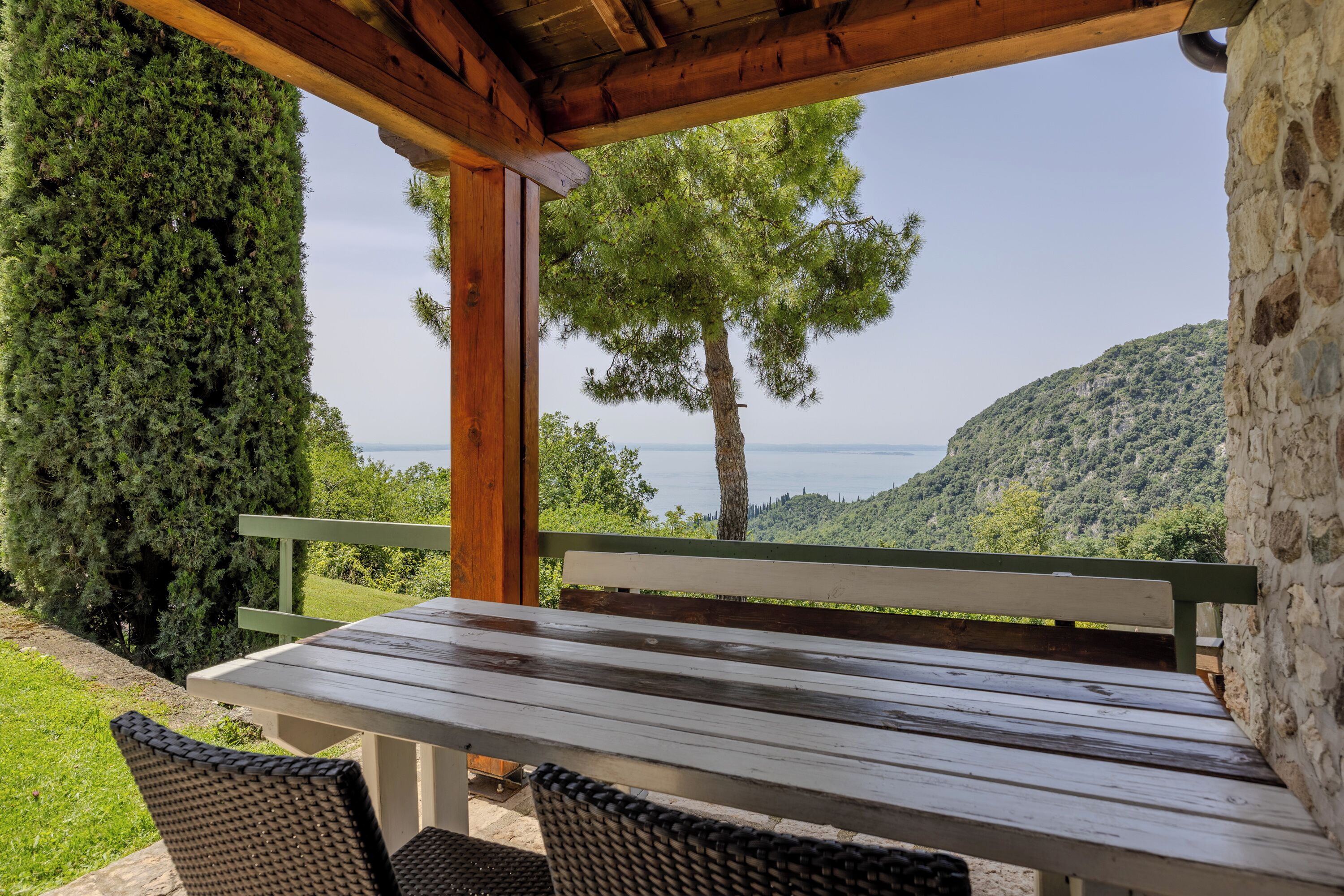 Een houten tafel en stoelen onder een pergola, met uitzicht op een boom en een wateroppervlak.